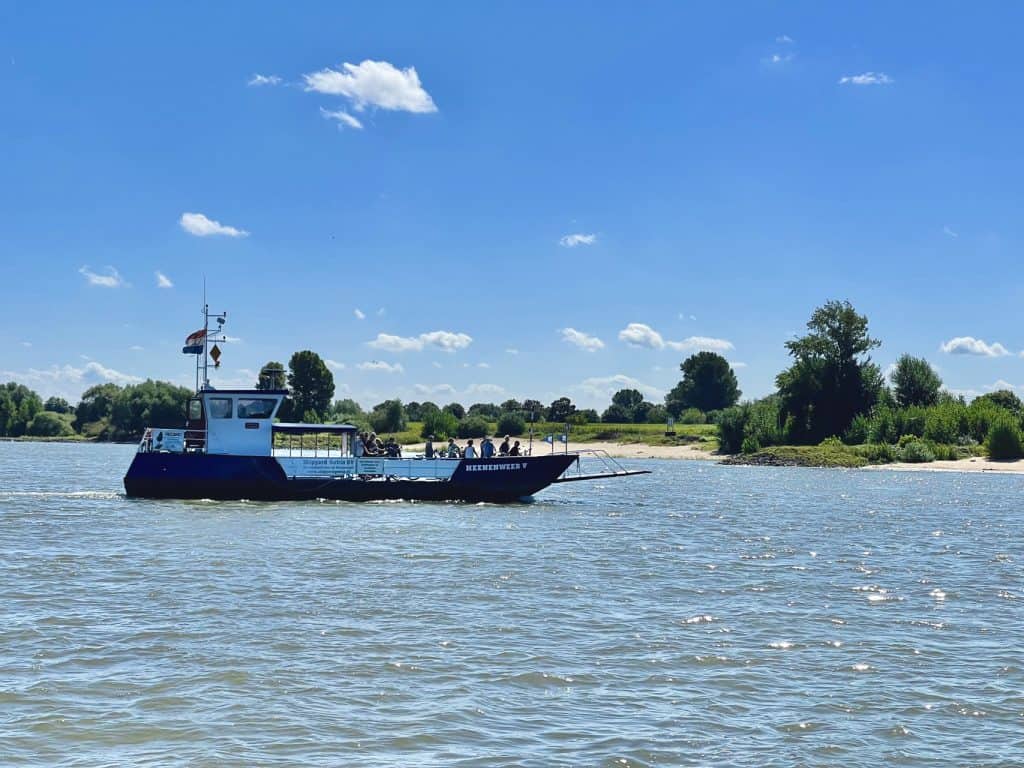 Zandmotor op de rivier bij 't Gelders Eiland, met een boot en groene oevers onder een heldere blauwe.