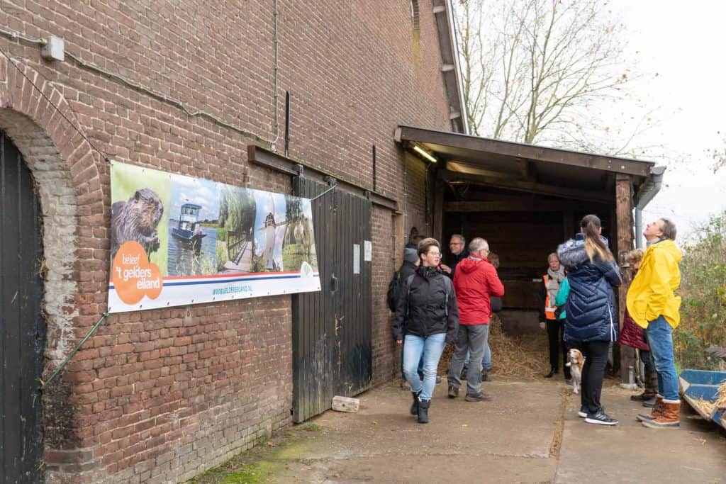 Kleine groep mensen bij de ingang van een boerderij op 't Gelders Eiland, met informatiebord over he.