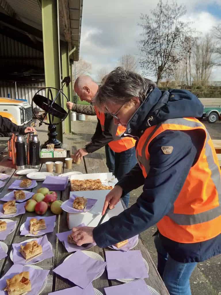 Verspreide mensen genieten van een buitenlunch met taart en fruit op 't Gelders Eiland, onder een be.