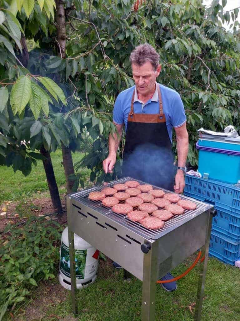 Gegrilde hamburgers op barbecue in tuin met groene bladeren en blauwe bakken op achtergrond.