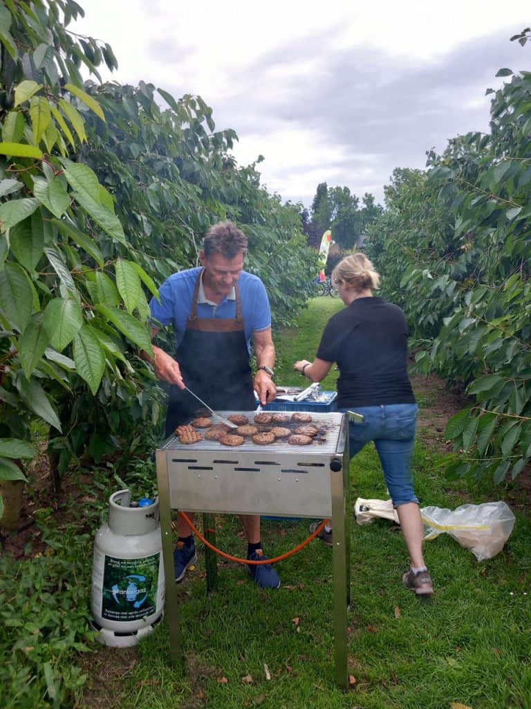 Houtskoolgrill met twee mensen die hamburgers bereiden op 't Gelders Eiland, omgeven door groene bom.