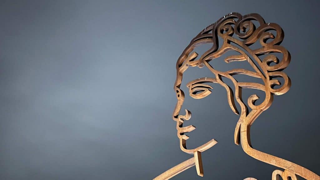 Houten kunstwerk van een vrouwenhoofd met sierlijke haarlokken, op een neutrale grijze achtergrond.