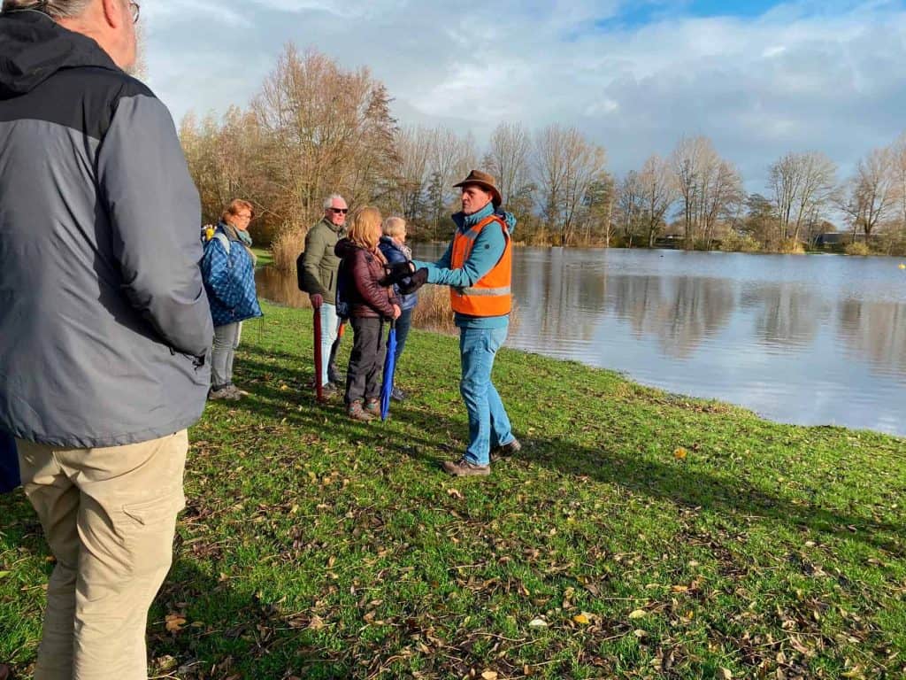 Een groep wandelaars ontvangt uitleg bij een rivier op 't Gelders Eiland, omgeven door bomen en een.