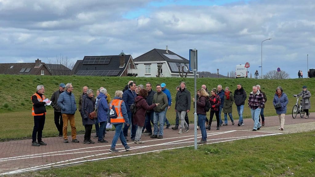 Gezelschap wandelaars op 't Gelders Eiland met huizen en een weg op de achtergrond.
