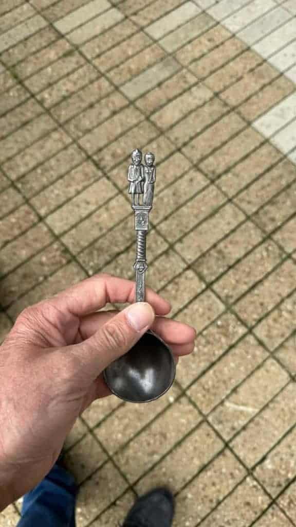 Kleine beeldhouwwerk op een lepel, vastgehouden door hand, op bestrate vloer.