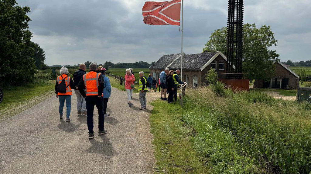 Kleine groep wandelaars bij 't Gelders Eiland met historische gebouwen en een vlag, in een landelijk.
