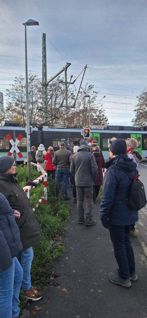 Mensen wachten bij overweg op 't Gelders Eiland, trein en spoorlijn zichtbaar.