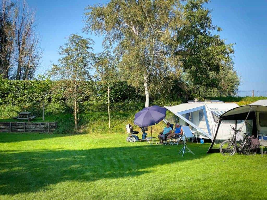 Kampeerplek met caravan, tent en zitje op grasveld in de natuur.