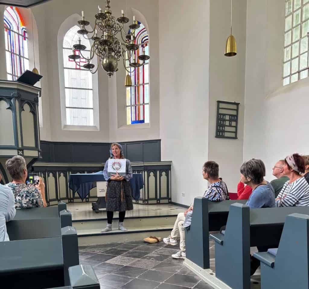 Een vrouw geeft een presentatie aan een kleine groep in een kerk met kleurrijke glas-in-loodramen. D.