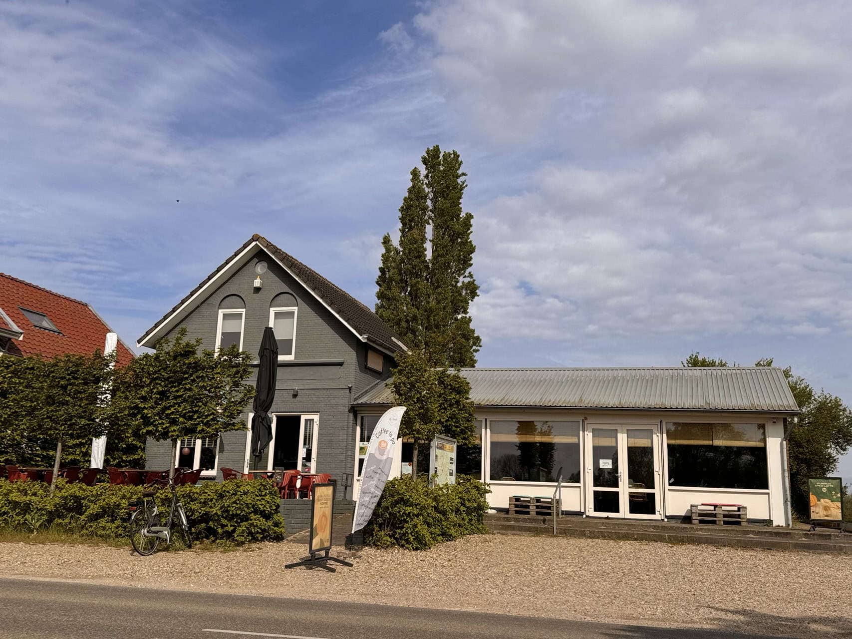 Gezellig café met terras en grote ramen op 't Gelders Eiland, omgeven door groen en een heldere blau.