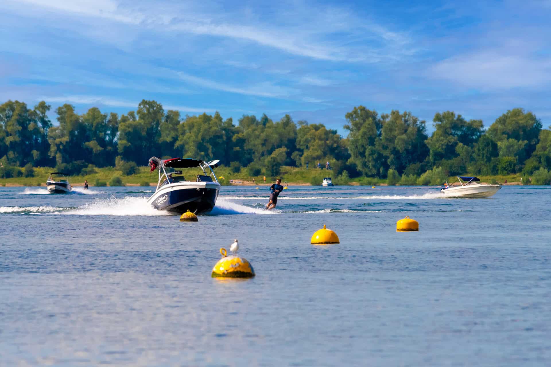 Zwemmer en boten op het water bij 't Gelders Eiland, met groene bomen en blauwe lucht op de achtergr.