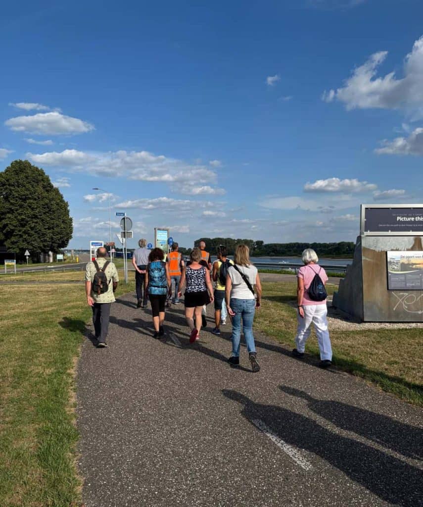 Gezelschap wandelaars op 't Gelders Eiland langs de rivier onder een heldere blauwe hemel.