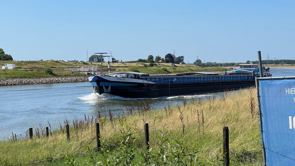 Vrachtboot vaart langs de oevers van 't Gelders Eiland onder een heldere blauwe hemel.