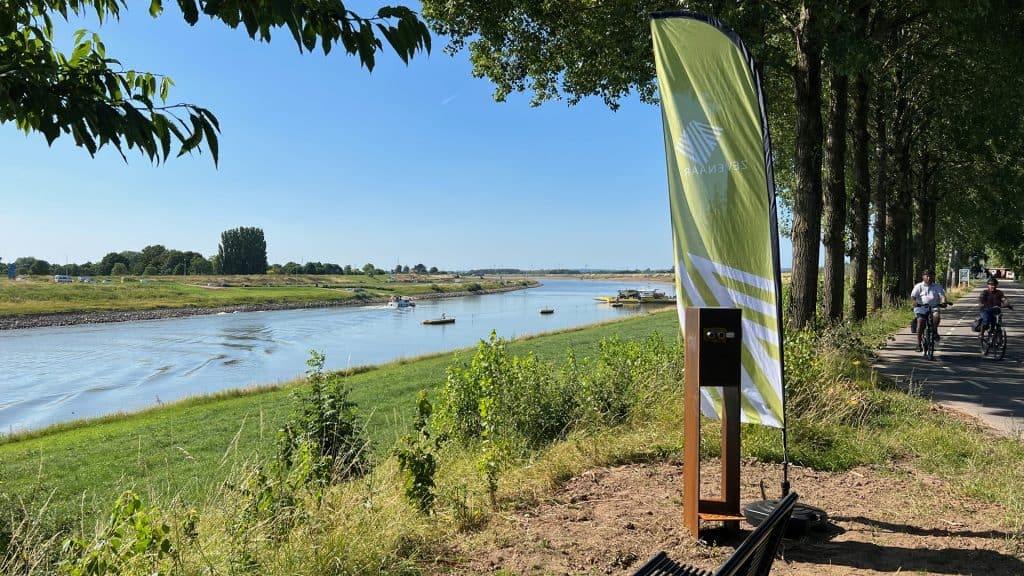 Fietsers langs de rivier op 't Gelders Eiland met groene bomen en blauwe lucht.