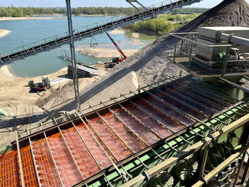 Zandwinning op 't Gelders Eiland met graafmachines en conveyorband, uitzicht op water en zandbergen.