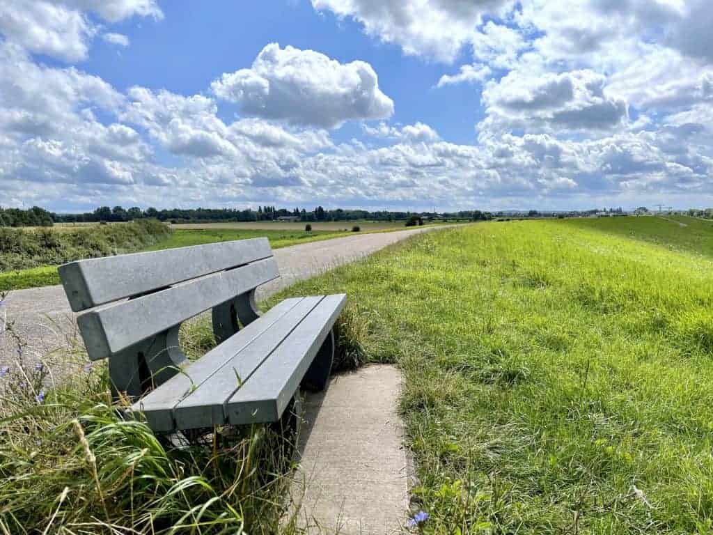 Rustieke bank langs wandelpad met uitzicht op groene velden en blauwe lucht.