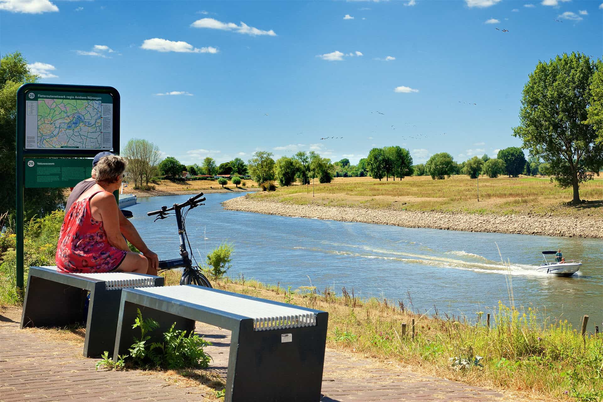 Vrouw zit op bankje bij rivier met fiets en informatiebord in 't Gelders Eiland.
