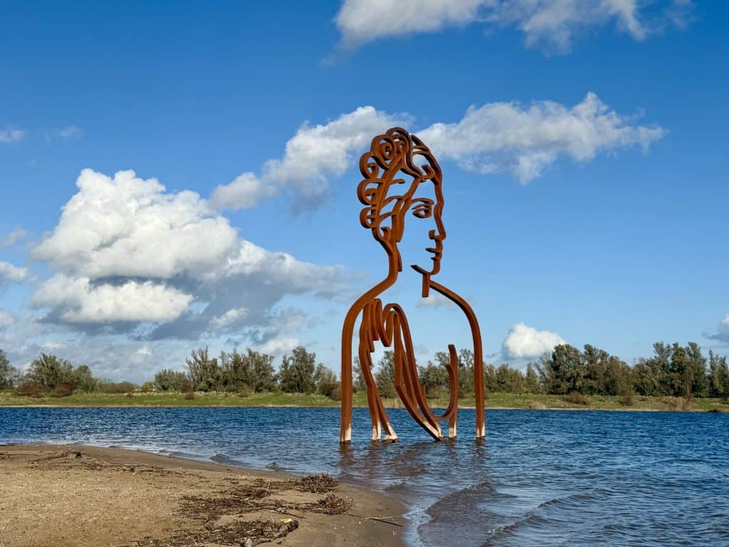 Kunstwerk van een grote metalen vrouwengestalte in het water op 't Gelders Eiland, met een heldere b.