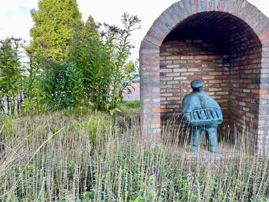Bronzen beeld onder een boog op 't Gelders Eiland, omgeven door gras en bomen.
