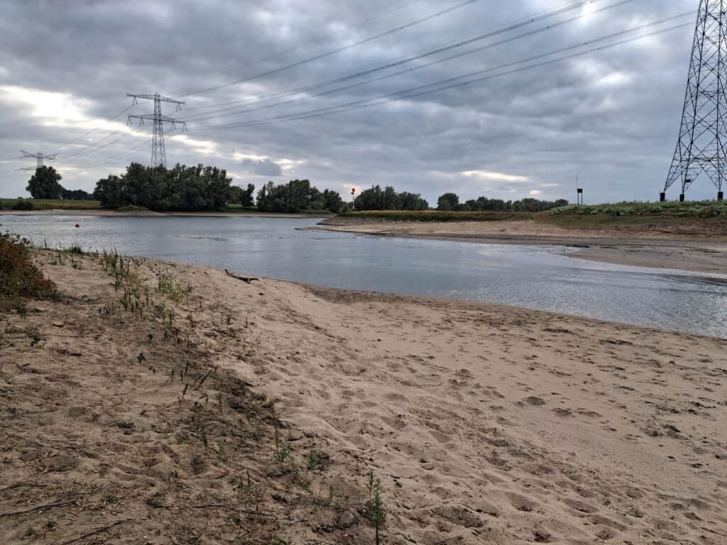 Rustige rivier met zandstrand en elektrische hoogspanningsmasten onder bewolkte hemel.
