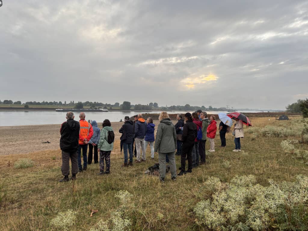 Groep mensen bij rivier op 't Gelders Eiland onder bewolkte hemel.